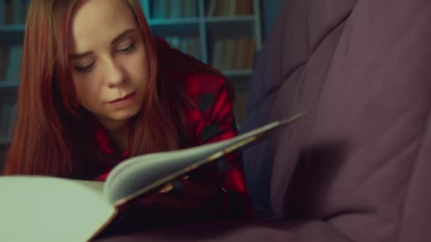 Een mooie student die een groot boek leest. Een attente jonge vrouw liggend op de bank met een boek en lezen in een donkere kamer in de avond — Stockvideo