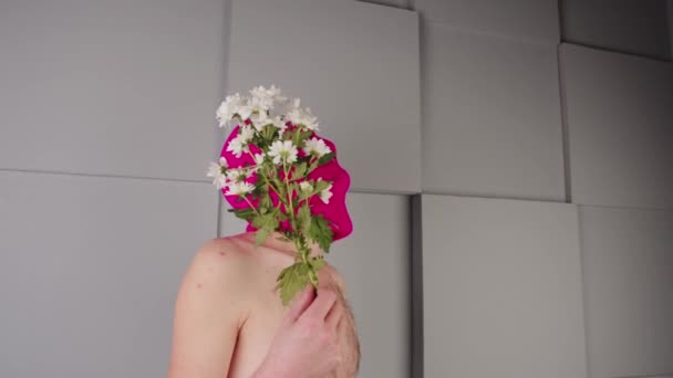 Shirless man in bivakmuts met bloemen in de buurt van grijs getextureerde geometrische muur. Onherkenbare man met naakte romp in roze bivakmuts die boeket madeliefjes vasthoudt en wegkijkt. — Stockvideo