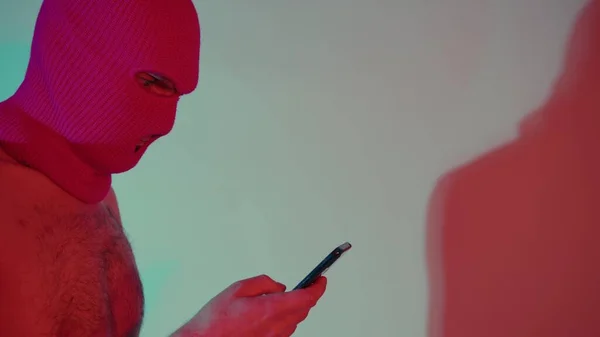 Anonymní muž bez trička v balaklavě prohledávající smartphone. Boční pohled na nebezpečného nerozpoznatelného muže s nahým trupem v balaklavě textové zprávy na moderním mobilním telefonu, když stojí v neonovém světle — Stock fotografie