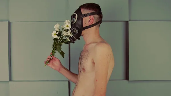 Muž bez trička v plynové masce, vonící květinami. Boční pohled na nerozpoznatelného samce s nahým trupem v černé plynové masce vonící kyticí čerstvých bílých květin u šedé stěny — Stock fotografie