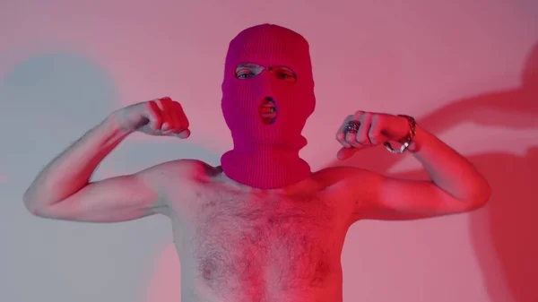 Anonymní muž bez trička v balaklávě ukazující biceps. Nerozpoznatelný muž s nahým trupem v růžové kuklě ukazující biceps a dívající se do kamery, zatímco stojí u stěny se stínem — Stock fotografie