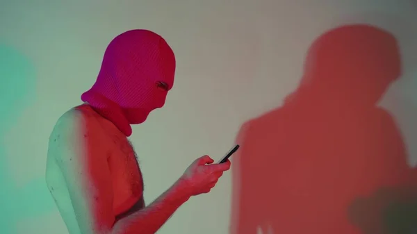 Anonymous shirtless άνθρωπος σε balaclava περιήγηση smartphone. Πλευρική άποψη του επικίνδυνου αγνώριστου άνδρα με γυμνό κορμό σε μήνυμα κειμένου balaclava στο σύγχρονο κινητό ενώ στέκεται στο φως νέον — Φωτογραφία Αρχείου