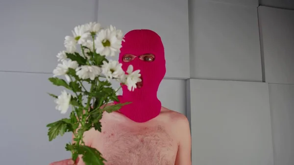 Névtelen félmeztelen férfi símaszkban virágokkal. Felismerhetetlen veszélyes férfi, meztelen törzzsel, rózsaszín maszkot visel, fehér virágokkal a kezében, szürke, texturált geometriai fal mellett. — Stock Fotó