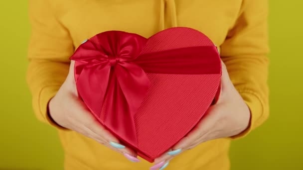 Parte del corpo della donna con scatola regalo rossa in forma di cuore su sfondo giallo. Persona irriconoscibile in possesso di regalo romantico con nastro. Concetto di presente durante giorno di San Valentino e giorno delle donne internazionale. — Video Stock