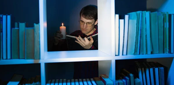 Mannen i glasögon med ljus läsa bok, står bakom bokhyllan. Man håller brinnande ljus medan du läser bok i trä bokhylla med olika litteratur i mörka rummet. — Stockfoto