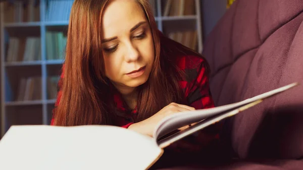 一个漂亮的学生在读一本大书 傍晚时分 一个心事重重的年轻女子躺在沙发上看书 在黑暗的房间里看书 免版税图库图片