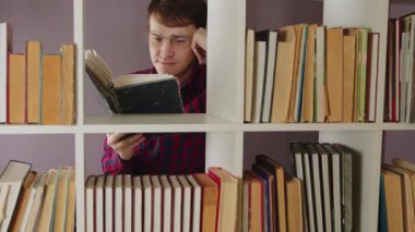Gözlüklü adam kitap okuyor, kitaplığın arkasında duruyor. Yorgun erkek, çeşitli edebiyat eserleriyle ahşap kitaplıkta kitap okurken gözlerini ovuşturuyor..