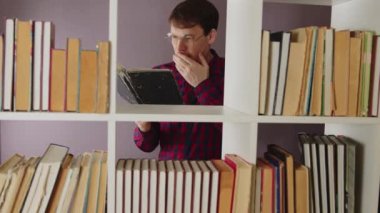 Kitaplığın yanında kitap okuyan ve dans etmeye başlayan bir adam. Gerçek zamanlı gözlüklü, esneyen, kitaplıktan kitap okumaktan sıkılan ve dans etmeye başlayan genç bir erkek.