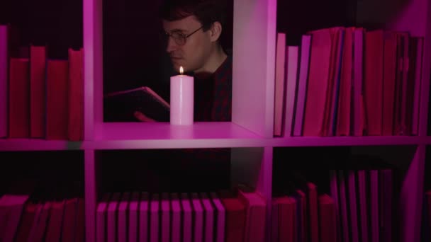 Serious man reading book near candle in bookcase. Male bookworm in eyeglasses reading book near burning candle placed on shelf of wooden bookcase with various literature in pink illumination — Stock Video