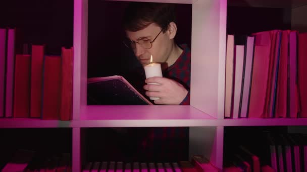 Mann in Gläsern mit Kerzenlesebuch, hinter Bücherregal stehend. Männchen hält brennende Kerze beim Lesen von Buch in Holzbücherregal mit diverser Literatur im dunklen Raum. — Stockvideo
