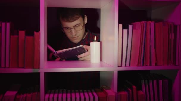 Homme sérieux lisant un livre près d'une bougie dans une bibliothèque. Ver de bibliothèque mâle dans les lunettes de lecture livre près de bougie brûlante placé sur l'étagère de la bibliothèque en bois avec diverses publications en éclairage rose — Video