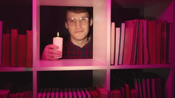 Mannen i glasögon med brinnande ljus står bakom bokhyllan. Ung man som håller vitt ljus i handen och tittar på kameran genom hyllan av bokhyllan. Porträtt av leende kille med olika litteratur. — Stockvideo