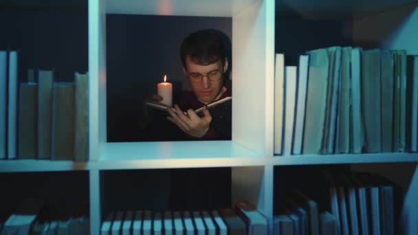 Man in glasses with candle reading book, standing behind bookshelf. Male holds burning candle while reading book in wooden bookcase with various literature in dark room. — Stock Video