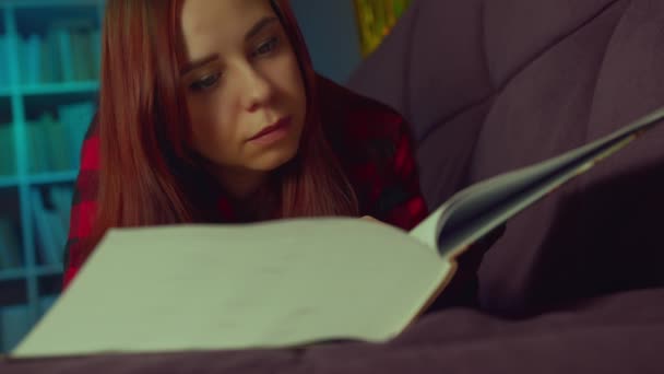 一个漂亮的学生在读一本大书。傍晚时分，一个心事重重的年轻女子躺在沙发上看书，在黑暗的房间里看书 — 图库视频影像