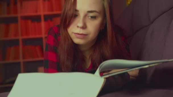 Una hermosa estudiante leyendo un gran libro. Una joven reflexiva tumbada en el sofá con un libro y leyendo en una habitación oscura por la noche — Vídeos de Stock