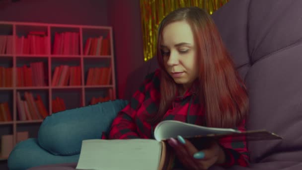 Гарний студент читає велику книжку. Вдумлива молода жінка лежить на дивані з книгою і читає в темній кімнаті ввечері — стокове відео
