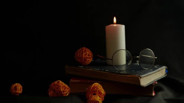 Καμένο κερί με γυαλιά που στέκονται πάνω σε παλιά βιβλία. Μεγάλο λευκό κερί καίει σε μαύρο φόντο που περιβάλλεται από διακόσμηση. Έννοια της ανάγνωσης σε σκοτεινό δωμάτιο στο φωτισμό της φλόγας. — Αρχείο Βίντεο