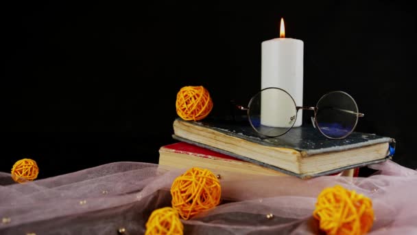 Libros de velas ardientes y velo delgado con decoración. Composición de vela de cera ardiente cerca de anteojos en pila de libros cerca de tela delgada con bolas de mimbre decorativas sobre fondo negro — Vídeo de stock