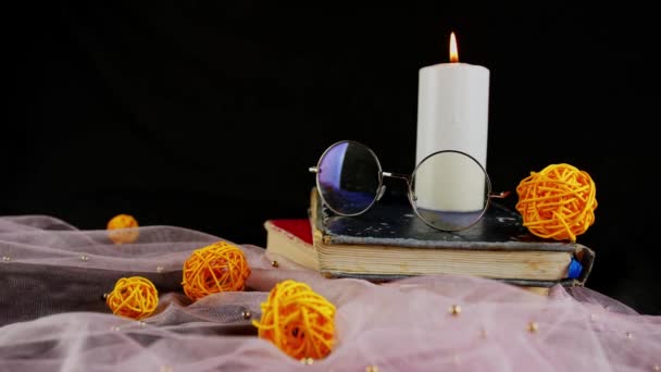 Спалювання свічок і тонка завіса з декором. Склад палаючої воскової свічки біля окулярів на стек книг біля тонкої тканини з декоративними плетеними кульками на чорному тлі — стокове відео