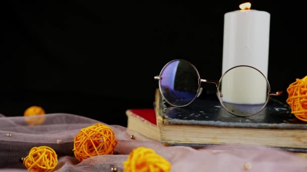 Libros de velas ardientes y velo delgado con decoración. Composición de vela de cera ardiente cerca de anteojos en pila de libros cerca de tela delgada con bolas de mimbre decorativas sobre fondo negro — Vídeos de Stock