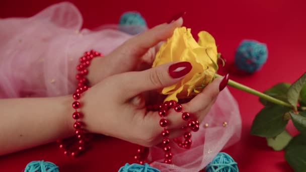 De dessus de la culture femme anonyme avec des perles rouges et avec manucure rouge tenant une rose jaune sur fond rouge en studio avec des boules décoratives en osier — Video