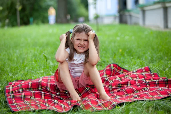 Little irritated girl preschooler sitting on plaid and grass — Stock Photo, Image