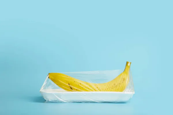 Frutas em embalagens plásticas do supermercado são mínimas. Banana em celofane e plástico não degradável sobre fundo azul. Embalagem de produtos biodegradáveis, protecção do ambiente, natureza — Fotografia de Stock