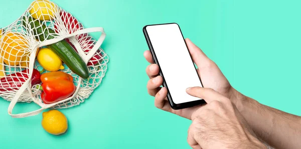 Grocery online background concept. A hand with a smartphone and eco-friendly bag with vegetables and fruit from the supermarket. Order, grocery and delivery food from eco-market. — Stock Photo, Image