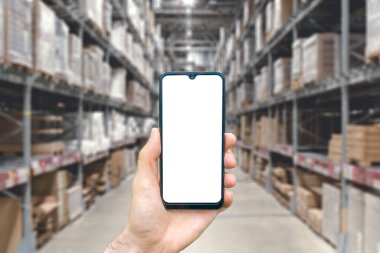Boş bir akıllı telefonu tutan eli olan bir depo. Ürün ve ürünlerle dolu bulanık bir sanayi deposu. Lojistik, iş, dağıtım, hizmet kavramı