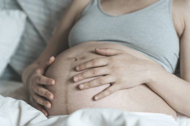 Hamile bir kadın, doğumdan önce karnını bir bebekle okşar güneşli, parlak bir yatak odasında yatarken. Kadın sağlığı, hamilelik, gebelik, gebelik kavramı.