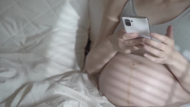 En gravid kvinde bruger telefonen, mens hun slapper af i en hjemmeseng. Brug af en smartphone før fødslen af et barn. Kvinders sundhed, fjernkonsultation med en læge, graviditetskoncept. – Stock-video