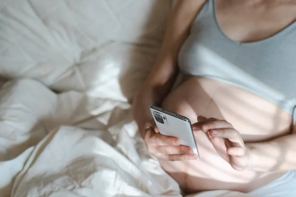 Eine schwangere Frau benutzt ein Smartphone, während sie es sich im heimischen Bett bequem macht. Internet, Einkaufen, Kommunikation während der Schwangerschaft und vor der Geburt eines Kindes. Gesundheit von Frauen und Neugeborenen — Stockfoto