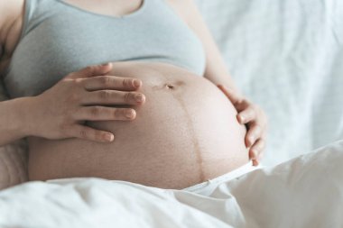 Hamile bir kadın, doğumdan önce karnını bir bebekle okşar güneşli, parlak bir yatak odasında yatarken. Kadın sağlığı, hamilelik, gebelik, gebelik kavramı.