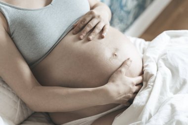 Hamile bir kadın, doğumdan önce karnını bir bebekle okşar güneşli, parlak bir yatak odasında yatarken. Kadın sağlığı, hamilelik, gebelik, gebelik kavramı.