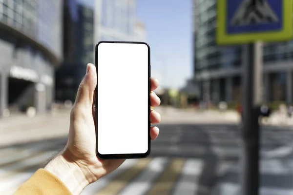 A smartphone in a hand with a blank screen against the backdrop of an urban street of the business quarter of the city. Business applications, transport, taxi, maps concept — Stock Photo, Image