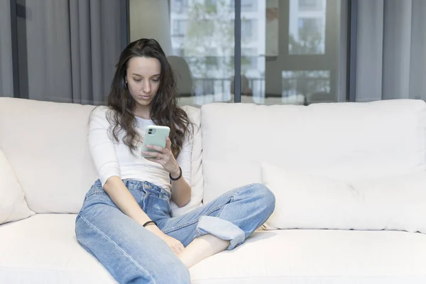 Junge Europäerin mit Smartphone in entspannter Umgebung zu Hause auf dem Sofa in der Freizeit. Internet, Technologiekonzept. — Stockfoto