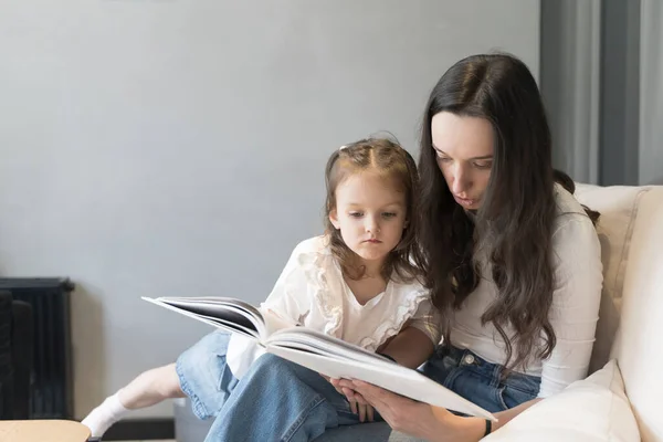 Mom and daughter are reading a book on the sofa. Study and activities with children at home, mental development, free time with children. High quality photo