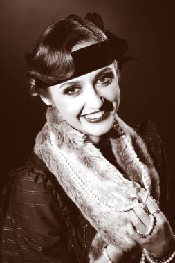 Mükemmel makyajlı, şaşaalı koyu renk saçlı bir mankenin portresi. Şık bir kolye, siyah bir kıyafet ve uzun bir bluz giyiyor. Ellerini başının arkasında tutuyor ve ileriye bakıyor..