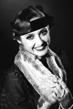 Mükemmel makyajlı, şaşaalı koyu renk saçlı bir mankenin portresi. Şık bir kolye, siyah bir kıyafet ve uzun bir bluz giyiyor. Ellerini başının arkasında tutuyor ve ileriye bakıyor..