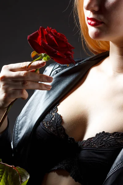 Portret młodej modelki w czerwonej sukience trzymającej kwiat róży w ręku nad ciemnym tłem w studiu fotograficznym. Koncepcja piękna i mody — Zdjęcie stockowe