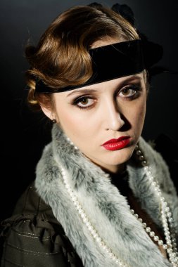 Mükemmel makyajlı, şaşaalı koyu renk saçlı bir mankenin portresi. Şık bir kolye, siyah bir kıyafet ve uzun bir bluz giyiyor. Ellerini başının arkasında tutuyor ve ileriye bakıyor..