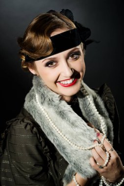 Mükemmel makyajlı, şaşaalı koyu renk saçlı bir mankenin portresi. Şık bir kolye, siyah bir kıyafet ve uzun bir bluz giyiyor. Ellerini başının arkasında tutuyor ve ileriye bakıyor..