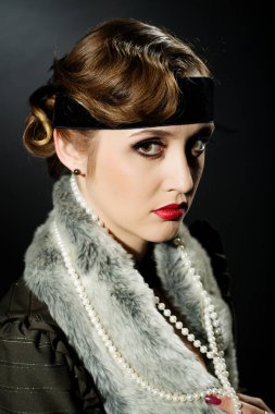 Mükemmel makyajlı, şaşaalı koyu renk saçlı bir mankenin portresi. Şık bir kolye, siyah bir kıyafet ve uzun bir bluz giyiyor. Ellerini başının arkasında tutuyor ve ileriye bakıyor..