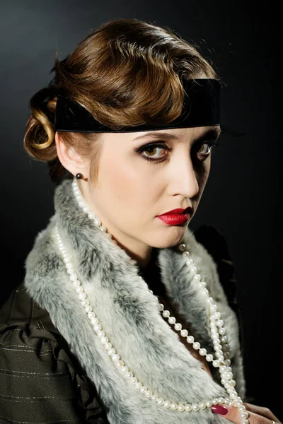 Mükemmel makyajlı, şaşaalı koyu renk saçlı bir mankenin portresi. Şık bir kolye, siyah bir kıyafet ve uzun bir bluz giyiyor. Ellerini başının arkasında tutuyor ve ileriye bakıyor..