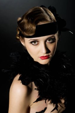 Mükemmel makyajlı, şaşaalı koyu renk saçlı bir mankenin portresi. Şık bir kolye, siyah bir kıyafet ve uzun bir bluz giyiyor. Ellerini başının arkasında tutuyor ve ileriye bakıyor..