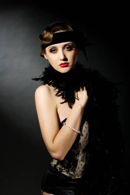 Mükemmel makyajlı, şaşaalı koyu renk saçlı bir mankenin portresi. Şık bir kolye, siyah bir kıyafet ve uzun bir bluz giyiyor. Ellerini başının arkasında tutuyor ve ileriye bakıyor..