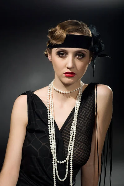 Portrait d'un modèle glamour aux cheveux foncés avec un maquillage parfait portant un collier luxueux, un body noir et un haut long, tenant ses mains derrière la tête et regardant droit devant. — Photo