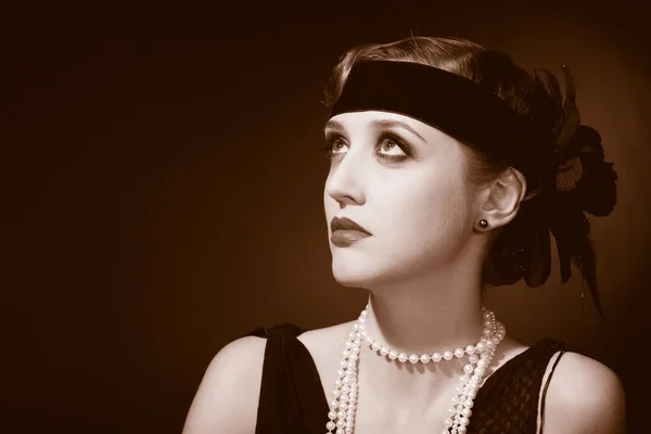 Mükemmel makyajlı, şaşaalı koyu renk saçlı bir mankenin portresi. Şık bir kolye, siyah bir kıyafet ve uzun bir bluz giyiyor. Ellerini başının arkasında tutuyor ve ileriye bakıyor..