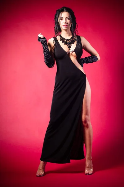 Siyah elbiseli ve çoraplı güzel bir kadın kırmızı arka planda kameraya poz veriyor.