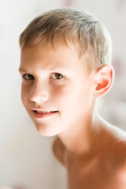 Sevimli, genç, neşeli, doğal bir gülüşü olan sarışın bir çocuk açık bej bir arka plana karşı çıplak omuzlarıyla mutlu bir şekilde gülümsüyor..